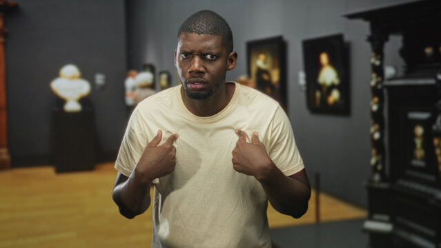 Man pointing thumbs to chest with questioning frown amid framed paintings in museum building, defensive posture and raised eyebrows; confusion doubt.