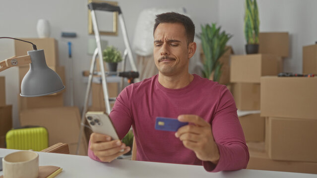 Young man with moustache holds smartphone and creditcard, frowning while checking a charge amid moving boxes in a building; financial frustration.