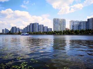 Urban waterfront scene with high-rise buildings, calm river reflections, and a passing boat under a bright blue sky with scattered clouds. © Ann