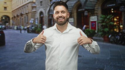 Hispanic man giving thumbs up, wearing white shirt on a cobblestone street by a historic stone...