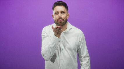 Obraz premium Man blowing a kiss with hand visible, smiling and wearing a white shirt against a purple studio backdrop; playful.
