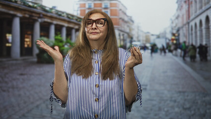 Fototapeta premium Woman holding glasses, shrugging with hands raised on a cobblestone street in a downtown shopping arcade; uncertainty decision dilemma.