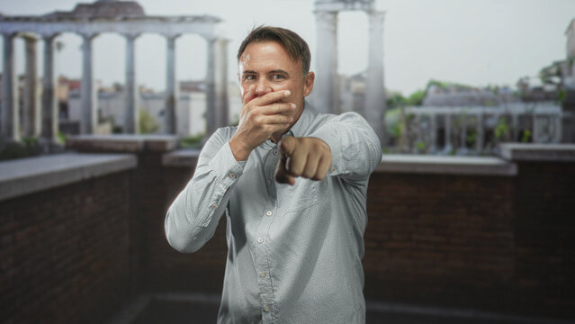 Man points finger at camera, laughs and alternately covers mouth while wearing a light patterned shirt on a building rooftop terrace with brick parapet and classical columns; playful confidence.