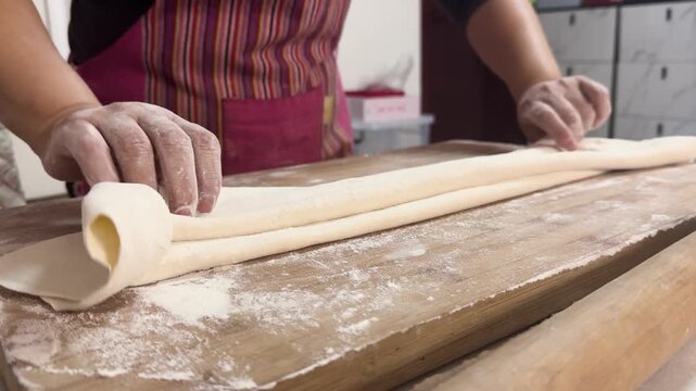 Preparing fresh noodles from scratch at home by shaping and cutting dough on a floured wooden board, showcasing traditional homemade cooking in a kitchen setting.