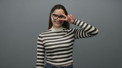 Fototapeta premium Woman wearing glasses and a striped turtleneck smiling and flashing a peace sign hand gesture in a gray studio; playful confidence.