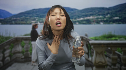 Fototapeta premium Young woman holding water bottle, hand at throat, coughing on a building terrace by a lake; discomfort recovery.
