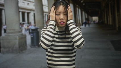 Woman covers mouth with hands in building colonnade, wearing black and white striped sweater, hoop earring and rings, then places hands on head in distress  anxiety isolation. © Krakenimages.com