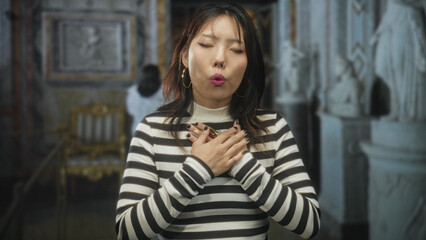 Fototapeta premium Woman with hands on chest and closed eyes in ornate museum building, striped sweater and visible rings; reverence.