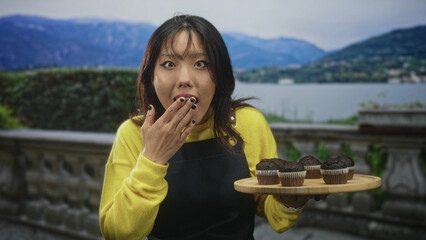 Young chinese woman holds wooden tray of chocolate muffins while hand covers mouth on building...