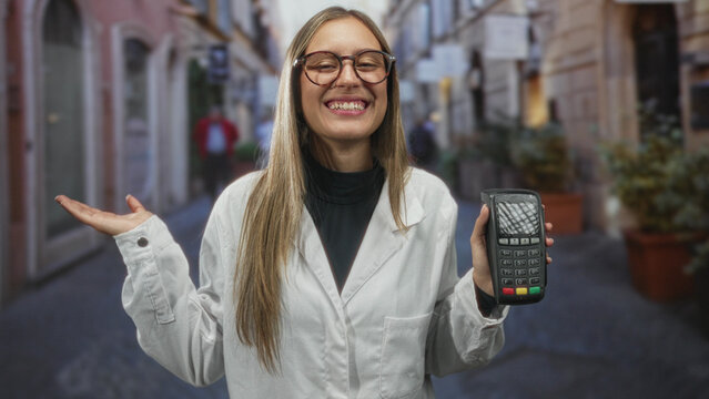 Woman in white coat holds card reader and gestures palms up on a narrow city street while presenting payment terminal; welcoming service.