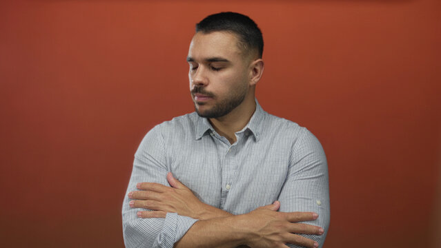 Hispanic man wearing striped shirt crosses arms in orange studio while closing eyes; rejection introspection.