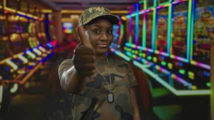 Fototapeta premium Young african american woman in army uniform shows thumbs up and thumbs down at casino slot machines under neon lights; decision making.