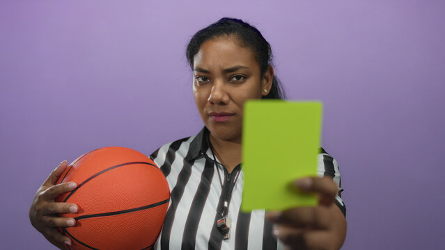 Referee woman in striped uniform holds basketball firmly and points yellow card with finger in studio; authority.