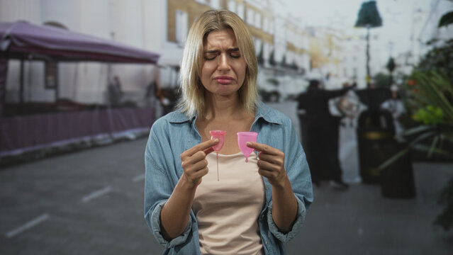 Woman holding two pink menstrual cups between fingers on a busy street, comparing small and large sizes with a displeased face; size comparison frustration.