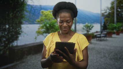 Obraz premium Woman in yellow dress holding a tablet, hands tapping screen and smiling while standing on a lakeside promenade with planters and a bench; serenity.
