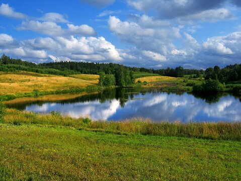 A peaceful lake surrounded by rolling green hills and meadows under a cloudy sky. Beautiful natural landscape of a Masurian lake at noon.