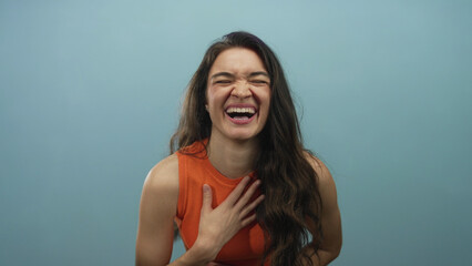 Fototapeta premium Woman laughs heartily with hand on chest and long brown hair falling over shoulders, wearing an orange sleeveless top against a light blue studio wall; joy.