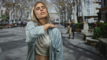 Fototapeta premium Woman holding neck in pain on busy city street with blurred pedestrians, benches, and bare trees; discomfort.