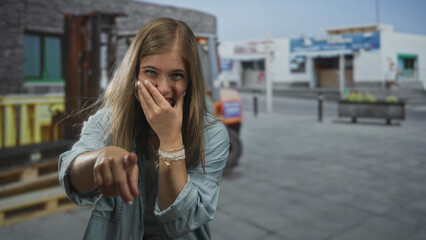 Fototapeta premium Young blonde woman points finger to camera while covering mouth in laughter on urban street; amusement.