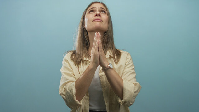 Woman with blonde hair praying with hands pressed together in studio against a light blue background; devotion.