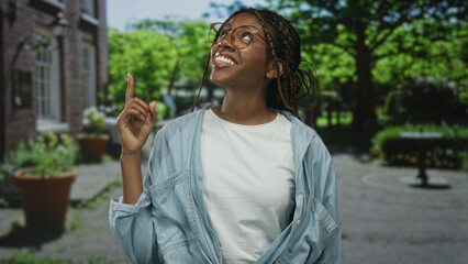 Fototapeta premium Woman points finger upward with a smiling face and glasses on a sunny street by a brick building and trees; optimism discovery.