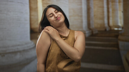 Naklejka premium Woman holding shoulder and wincing in front of a stone building colonnade wearing brown tanktop; discomfort.