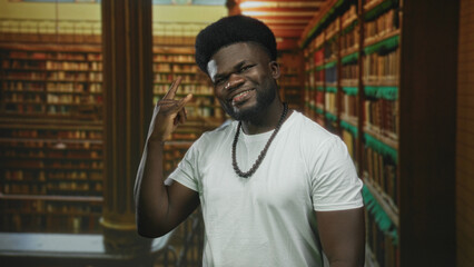 Man showing rock horns with right hand and smiling amid tall bookshelves in a library building aisle  confidence study focus. © Krakenimages.com