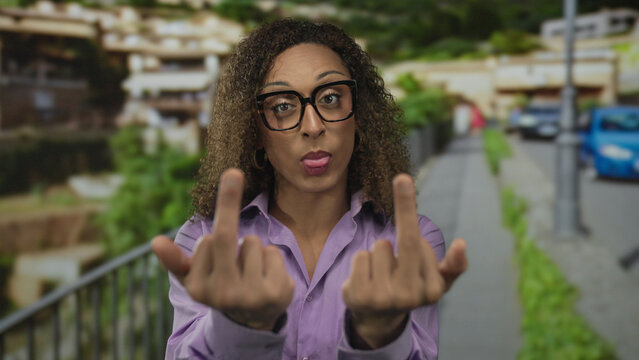 Trans woman raising middle finger at camera on street near sidewalk railing amid blurred city buildings; defiance.