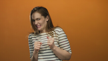 Fototapeta premium Woman grimacing and clenching fists with raised hands in orange studio wearing striped shirt; disgust.