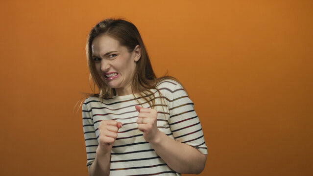 Woman grimacing and clenching fists with raised hands in orange studio wearing striped shirt; disgust.