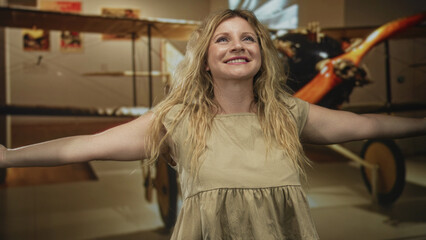 Blonde woman smiling with arms outstretched in front of a vintage propeller plane inside a museum building wearing a tan dress  joyful wonder exploration curiosity. © Krakenimages.com