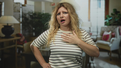 Fototapeta premium Woman clutch chest, mouth open, blurry living room with lamps and armchair, blonde in a striped shirt inside a building; distress.