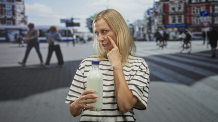 Fototapeta premium Woman holding milk bottle points finger to cheek in busy urban street with tram and cyclists nearby; contemplation curiosity.