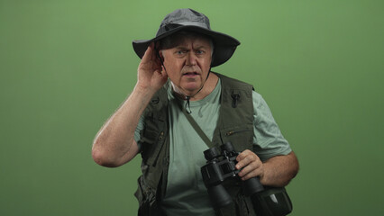 Fototapeta premium Senior man with binoculars cupping ear and listening, wearing fishing hat and field vest in studio; curiosity exploration.
