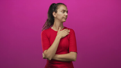 Obraz premium Young hispanic woman finger to chin, arms crossed, red top, ponytail, pink backdrop in studio; thoughtful reflection.