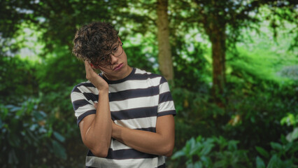 Fototapeta premium Teenage man with glasses resting hand on cheek, arms crossed, standing in forest with dense green foliage; quiet pensive.