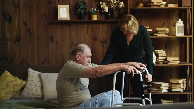Caring middle-aged woman helps her weak senior dad to use a walking frame
