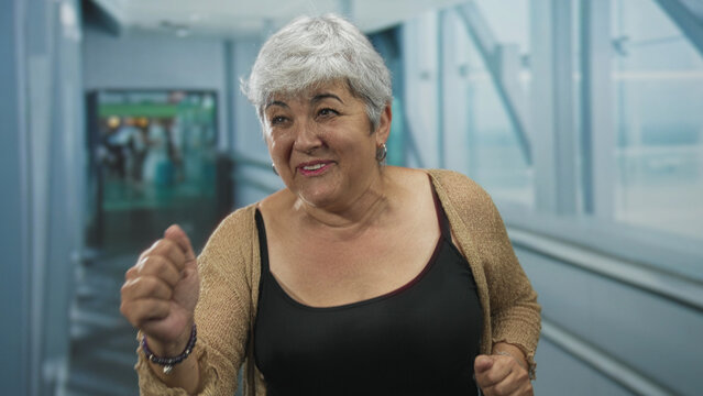Woman raises fists in celebration on airport moving walkway while smiling and walking toward a gate; joy travel triumph.