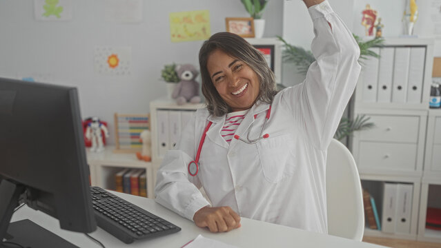 Hispanic woman doctor in white coat with stethoscope raising arm and smiling at desk with computer and pediatric toys in building; joy celebration.