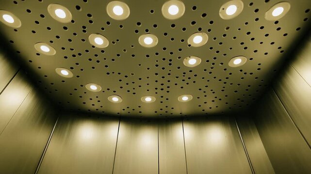 Ascending inside a modern elevator cabin featuring a detailed ceiling with numerous bright recessed lights and a dark perforated panel design, creating a contemporary indoor atmosphere