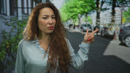 Woman points finger to the right on street wearing light blue shirt with skeptical expression;...