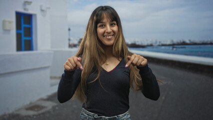Obraz premium Woman points finger to camera on street by white building and seaside promenade with blue window and railing; joyful playful vacation.