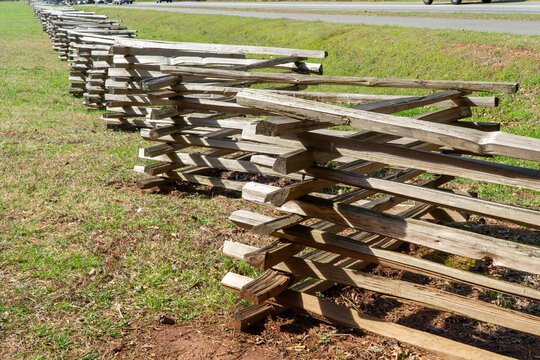 Weathered, rustic wooden split rail fence, also known as a zigzag or snake fence, stretching across a vibrant green field with a distant rural road under a bright daytime sky.