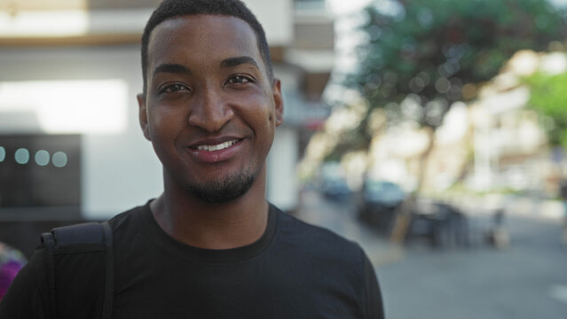Young black man smiling with backpack strap on shoulder, short beard and dimples, looking at camera in street near cafe and building facades; confidence.