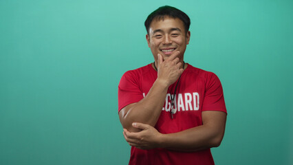 Fototapeta premium Young chinese lifeguard man in red shirt with lifeguard print and whistle holds chin in studio with green wall; contemplation.