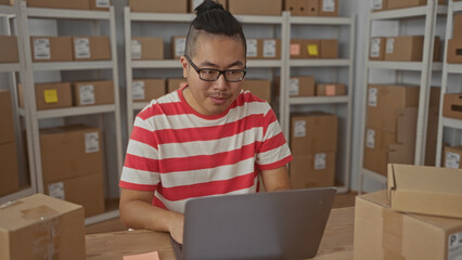 Man in striped shirt typing on laptop among parcel boxes with a fist pump gesture in storage...