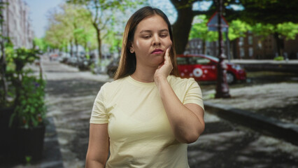 Fototapeta premium Young woman with hand on cheek for toothache on city street near parked car and tree lined sidewalk; discomfort.