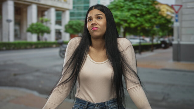 Woman shrugging shoulders on busy street beside commercial building with raised arms and puzzled expression; confusion.