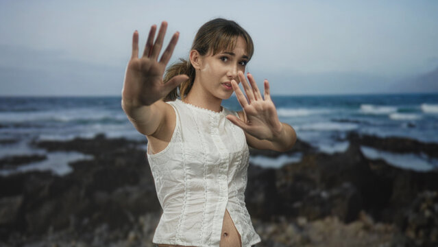 Young woman with hands raised to block camera at rocky seaside, white sleeveless blouse and palms forward; privacy solitude.
