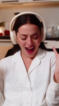 Young woman at night home wears headphones hears song starts seated dance laughs cheers yeah. Caucasian girl on sofa at evening makes rock hand signs nods to beat smiles shows joy fun energy music oh.